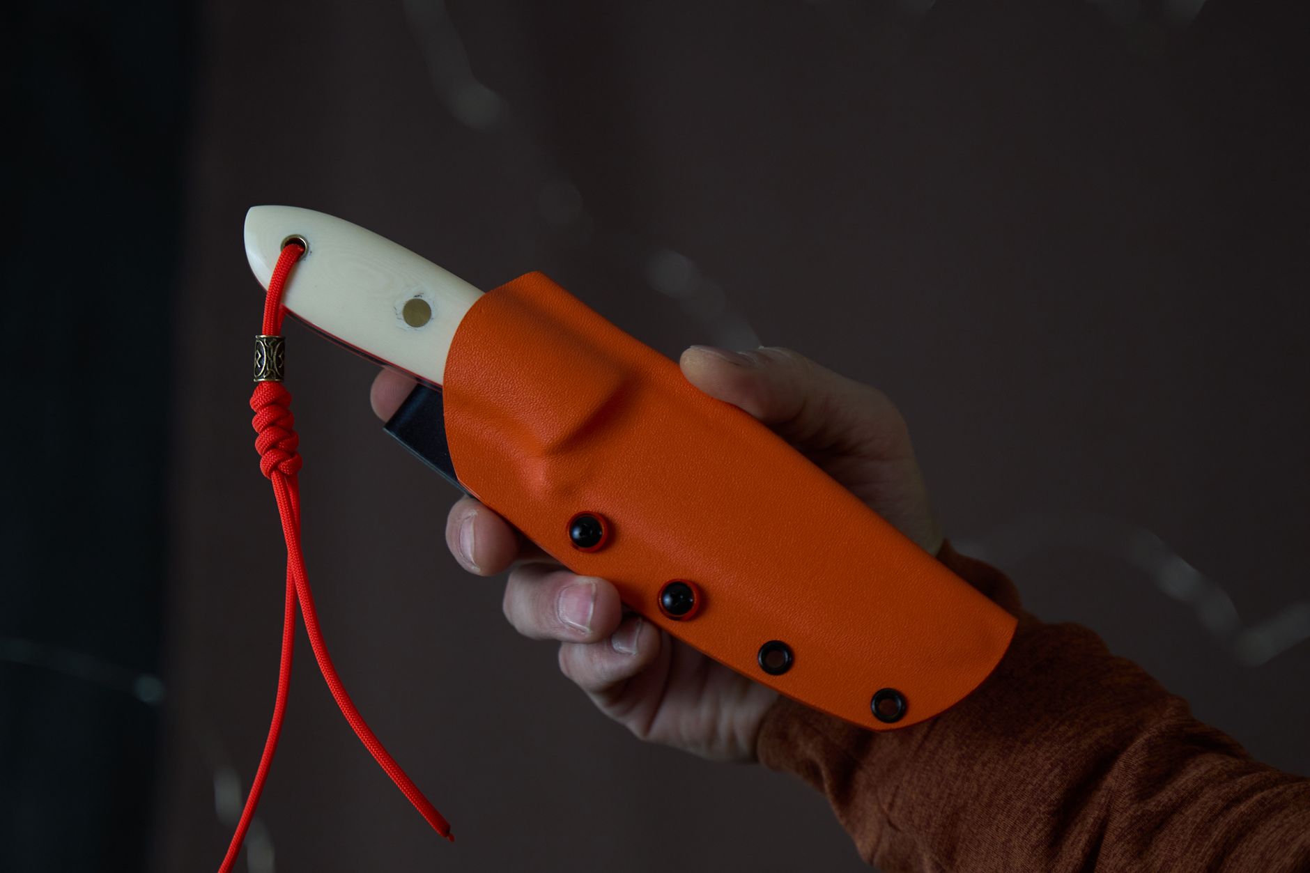 A person holds a knife with an orange sheath and white handle, featuring a red cord.