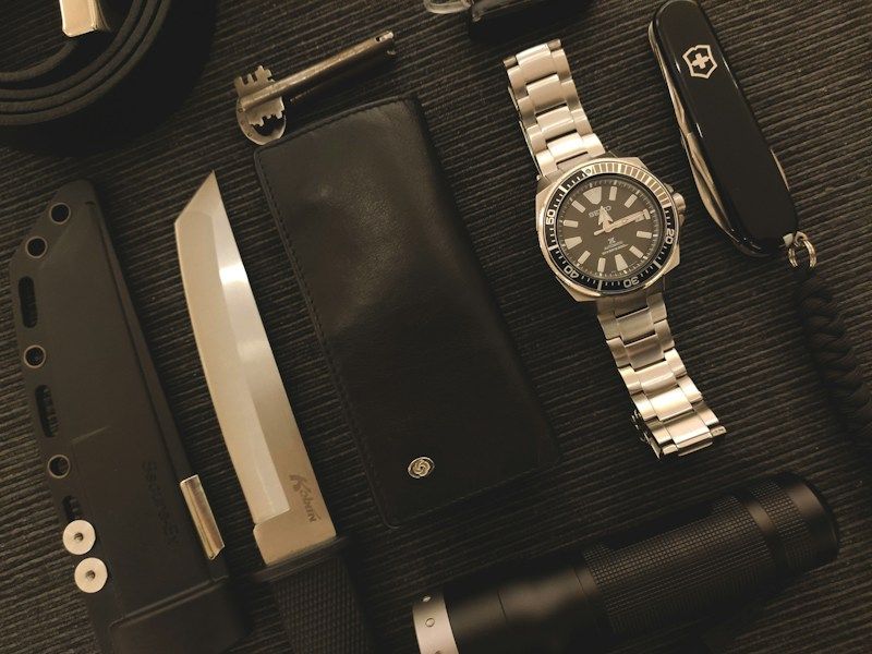 Everyday carry flatlay with watch, knife, wallet, and flashlight on dark surface